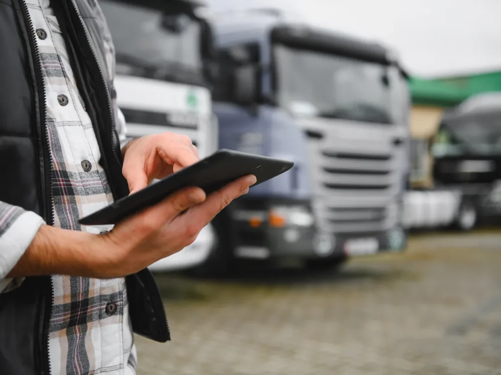 Worker in a plaid shirt using a tablet in front of parked semi trucks, illustrating on-site physical security risk assessments
