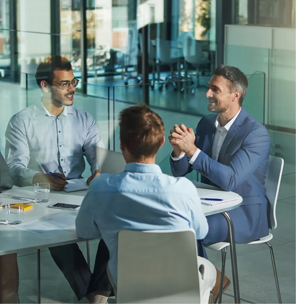 Three colleagues talking around a table in a glass-walled meeting room, illustrating compliance frameworks in practice